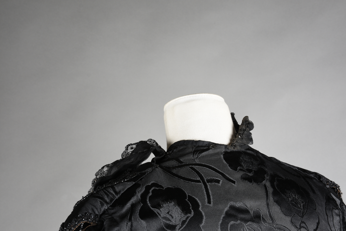 Kjoleliv av sort brokade. Silkeinnfelling og tyllinnfelling foran. Stjernekrave besatt med slag av paljetter og perler. Høy halslinning. Brokaden har et valmuemønster (jugend).