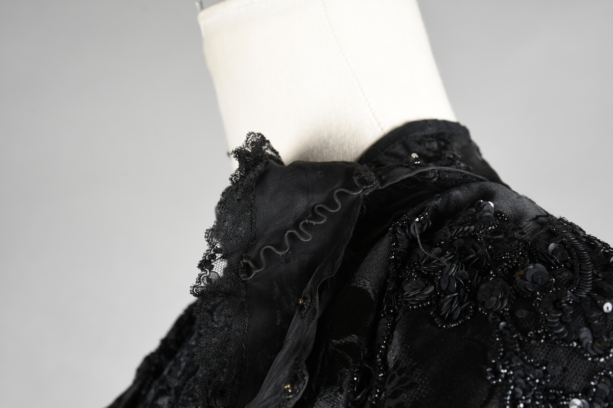 Kjoleliv av sort brokade. Silkeinnfelling og tyllinnfelling foran. Stjernekrave besatt med slag av paljetter og perler. Høy halslinning. Brokaden har et valmuemønster (jugend).