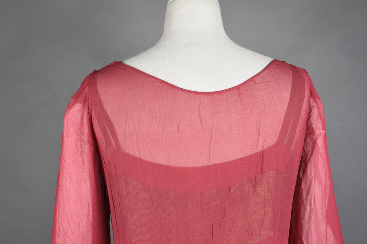 Overkjole av bringebærrød chiffon med garnityr av bronsebrun taft lagt på som skjell og med tre-motiver i tykksømsstrek. I sidene og midt foran oppheftet med bronsebånd (metalltråd?). Langs kanten nede og på ermene er den pyntet med et bredt stykke av brune silkespisser. Halvlange ermer, dyp utringning. Strikk nederst på ermene.