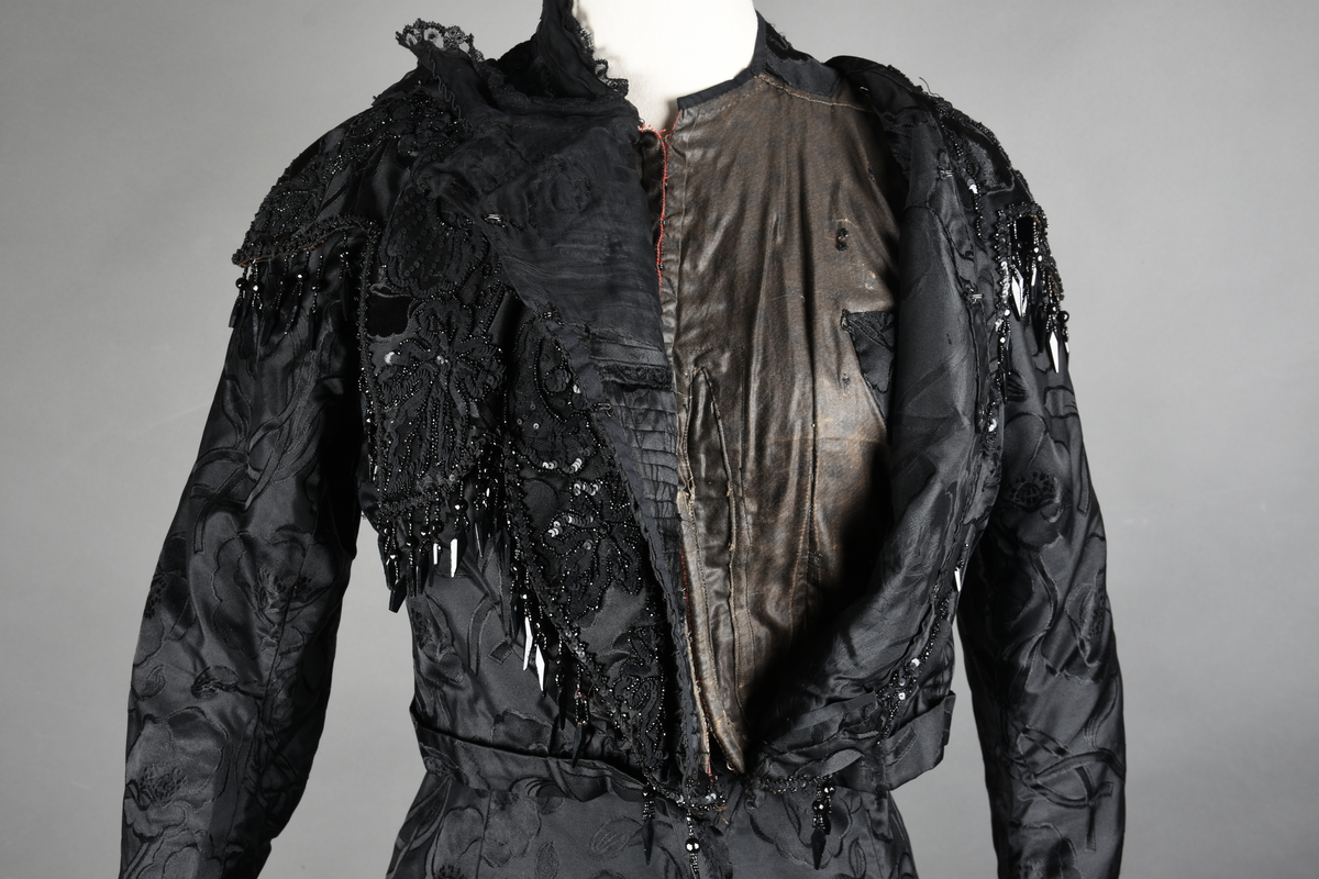 Kjoleliv av sort brokade. Silkeinnfelling og tyllinnfelling foran. Stjernekrave besatt med slag av paljetter og perler. Høy halslinning. Brokaden har et valmuemønster (jugend).