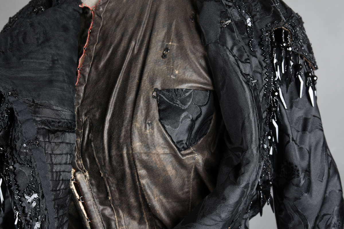 Kjoleliv av sort brokade. Silkeinnfelling og tyllinnfelling foran. Stjernekrave besatt med slag av paljetter og perler. Høy halslinning. Brokaden har et valmuemønster (jugend).