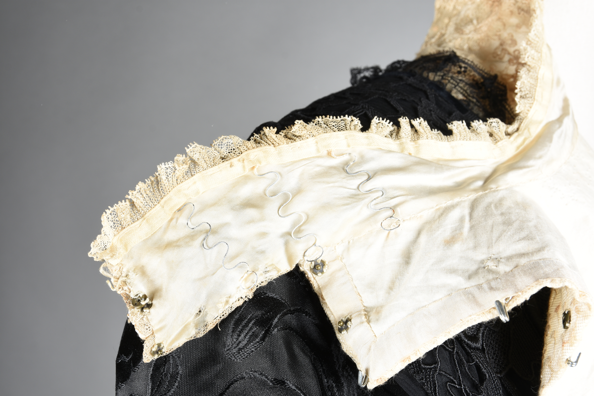 Kjoleliv av brokade med valmuemønster (jugend). Hvit innfelling foran, og rundt mansjettene. Hvitt tyllbroderi. Sjal og ermegarnityre av kraftig sort blonde på chiffonbunn. Sort fløyelslivbånd med tre ender. Høy halslinning.