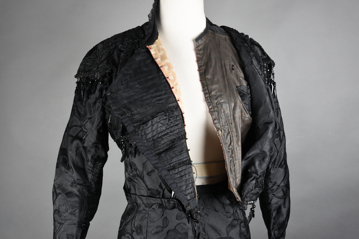 Kjoleliv av sort brokade. Silkeinnfelling og tyllinnfelling foran. Stjernekrave besatt med slag av paljetter og perler. Høy halslinning. Brokaden har et valmuemønster (jugend).