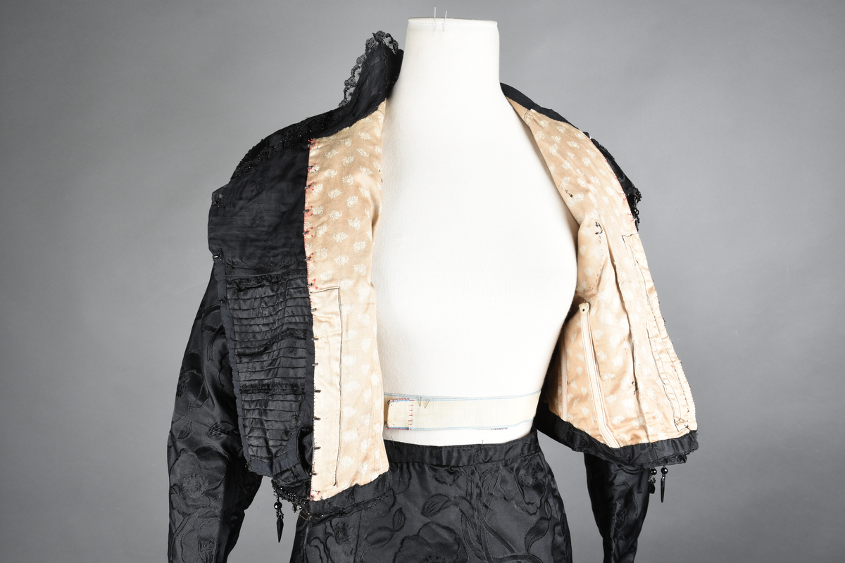 Kjoleliv av sort brokade. Silkeinnfelling og tyllinnfelling foran. Stjernekrave besatt med slag av paljetter og perler. Høy halslinning. Brokaden har et valmuemønster (jugend).