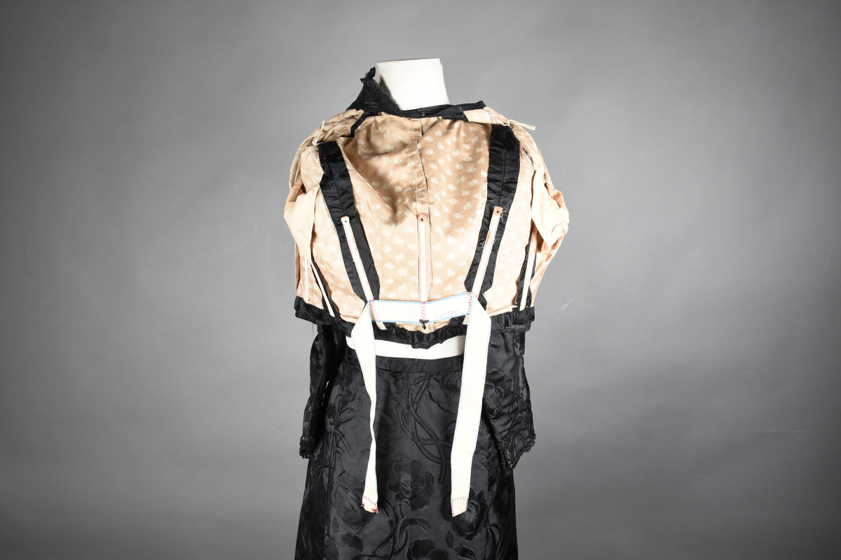 Kjoleliv av sort brokade. Silkeinnfelling og tyllinnfelling foran. Stjernekrave besatt med slag av paljetter og perler. Høy halslinning. Brokaden har et valmuemønster (jugend).