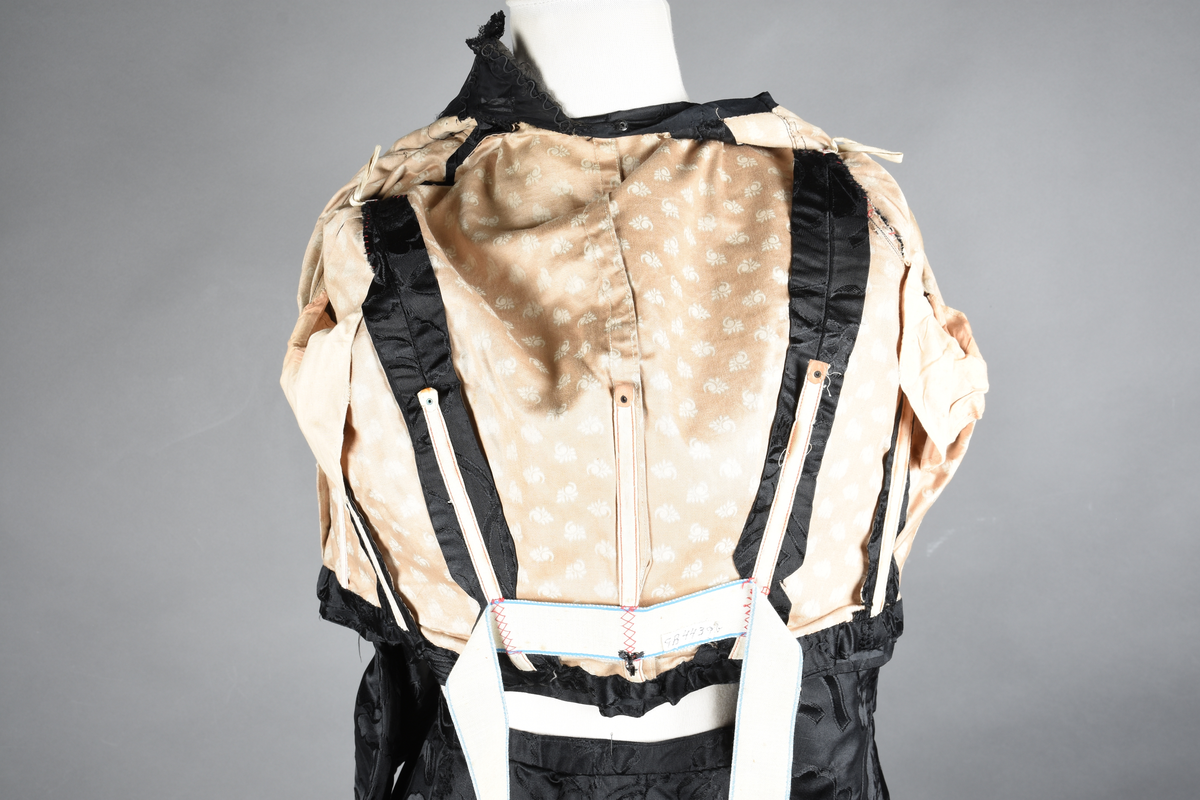 Kjoleliv av sort brokade. Silkeinnfelling og tyllinnfelling foran. Stjernekrave besatt med slag av paljetter og perler. Høy halslinning. Brokaden har et valmuemønster (jugend).