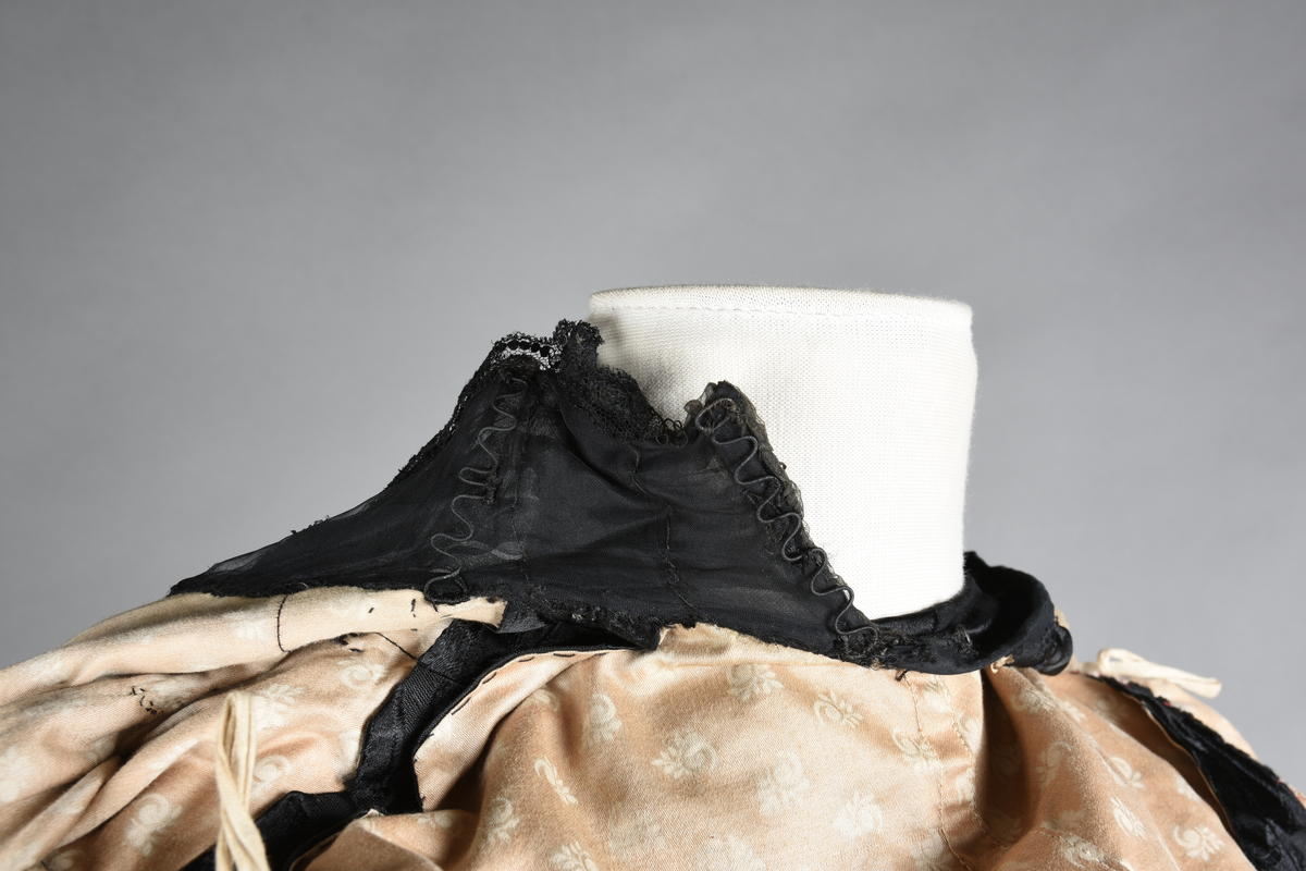 Kjoleliv av sort brokade. Silkeinnfelling og tyllinnfelling foran. Stjernekrave besatt med slag av paljetter og perler. Høy halslinning. Brokaden har et valmuemønster (jugend).