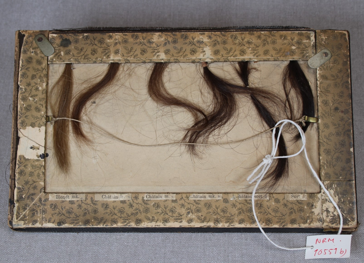 Tolv hårprøver i ulike fargenyanser festettil to rektangulære plansjer i tykk papp. En merkelapp under hver prøve viser fargens betegnelse.