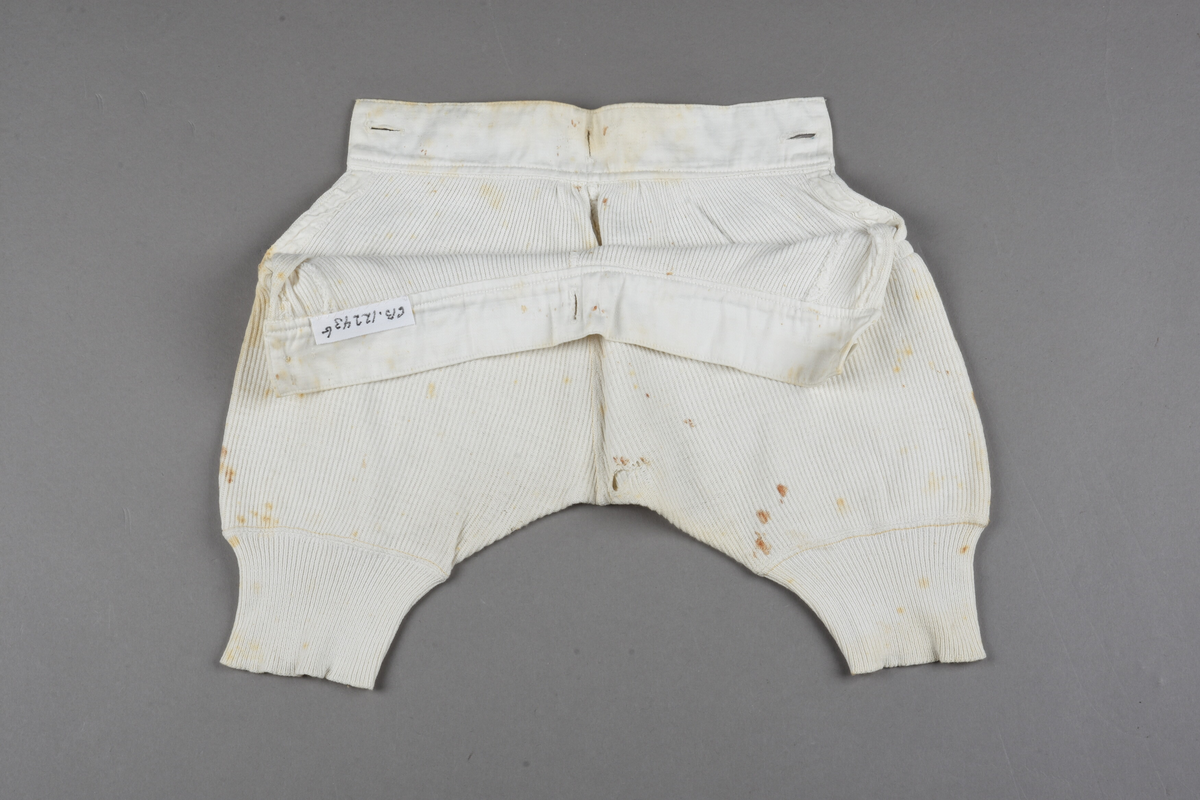 Underbenklær til barn av stripet hvit makó. Splitt på begge sider, knappes i begge sider av linningen. Tekstiltrekte knapper. Linningen er forsterket med lerret foran. Ekstra knapphull midt på, bak på linningen. Trangere nederst på beina for å holdes på plass. Rustflekker.