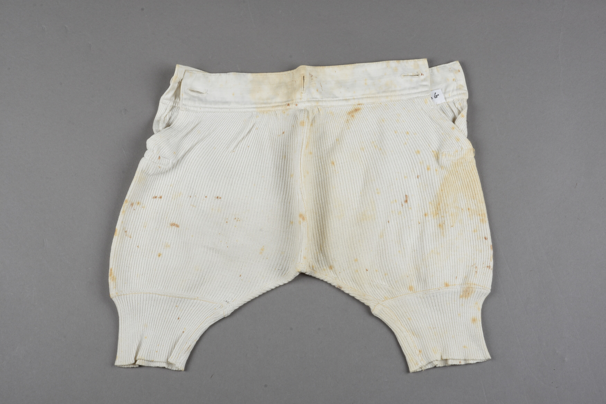 Underbenklær til barn av stripet hvit makó. Splitt på begge sider, knappes i begge sider av linningen. Tekstiltrekte knapper. Linningen er forsterket med lerret foran. Ekstra knapphull midt på, bak på linningen. Trangere nederst på beina for å holdes på plass. Rustflekker.