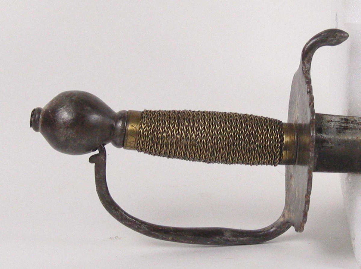 Soldatkorde av type M1701. Pæreformet knapp med nitteknapp. Fliket nyreformet parerplate med nedbøyd bakre parerstang. Fremre parerstang går over i håndbøylen som er tappet inn i knappen. Grepet har snodd messingtråd som avluttes med messingbånd oppe og nede. Rett tveegget klinge med midtås og rester av Fredrik IVs monogram.