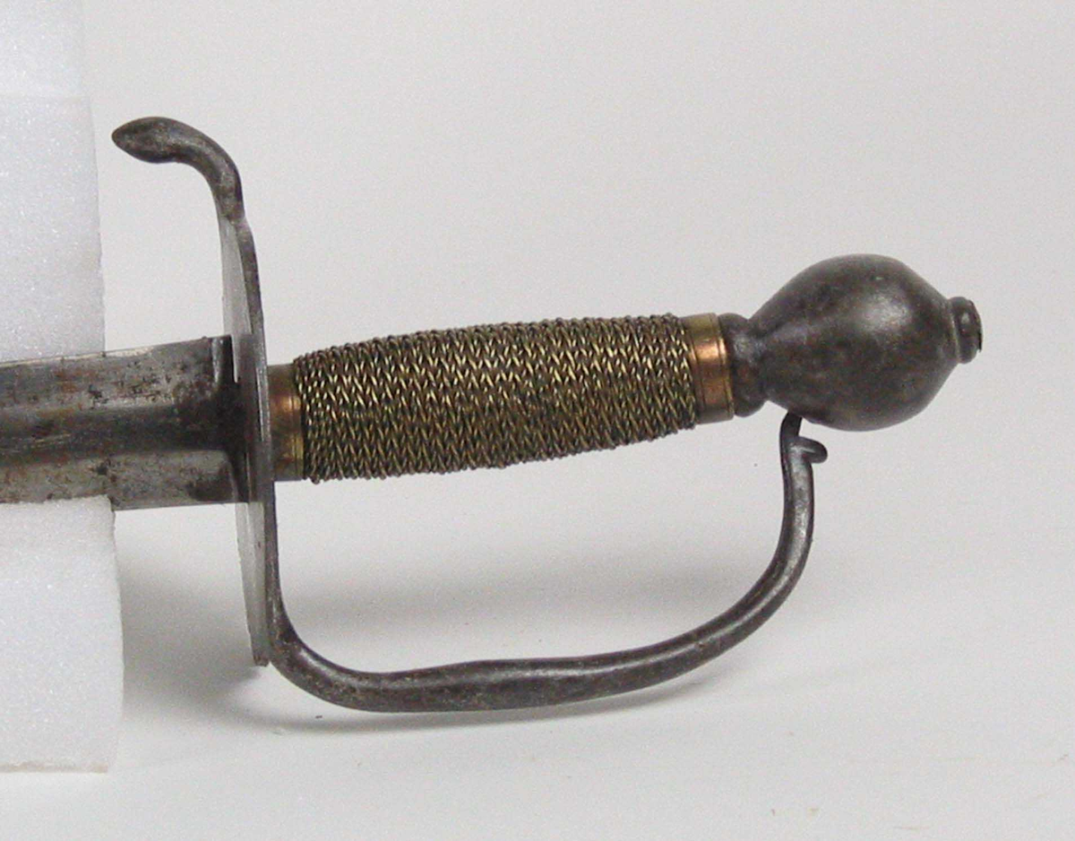 Soldatkorde av type M1701. Pæreformet knapp med nitteknapp. Fliket nyreformet parerplate med nedbøyd bakre parerstang. Fremre parerstang går over i håndbøylen som er tappet inn i knappen. Grepet har snodd messingtråd som avluttes med messingbånd oppe og nede. Rett tveegget klinge med midtås og rester av Fredrik IVs monogram.