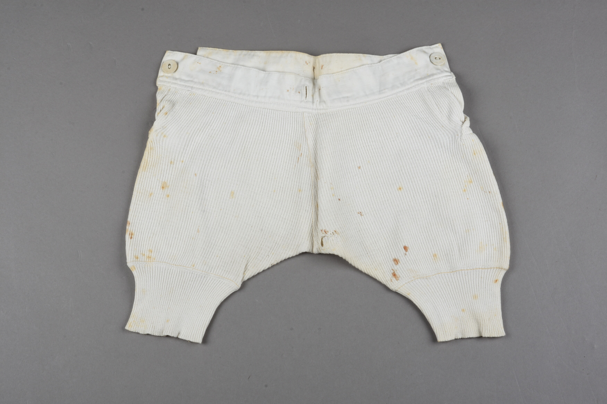 Underbenklær til barn av stripet hvit makó. Splitt på begge sider, knappes i begge sider av linningen. Tekstiltrekte knapper. Linningen er forsterket med lerret foran. Ekstra knapphull midt på, bak på linningen. Trangere nederst på beina for å holdes på plass. Rustflekker.