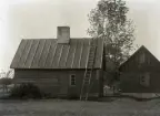 Här ser vi stugans baksida. Bakdörren tyder på att det fanns ett litet kök innanför framför skorstensstocken. T h måste bostadsrummet ha legat och t h var det 