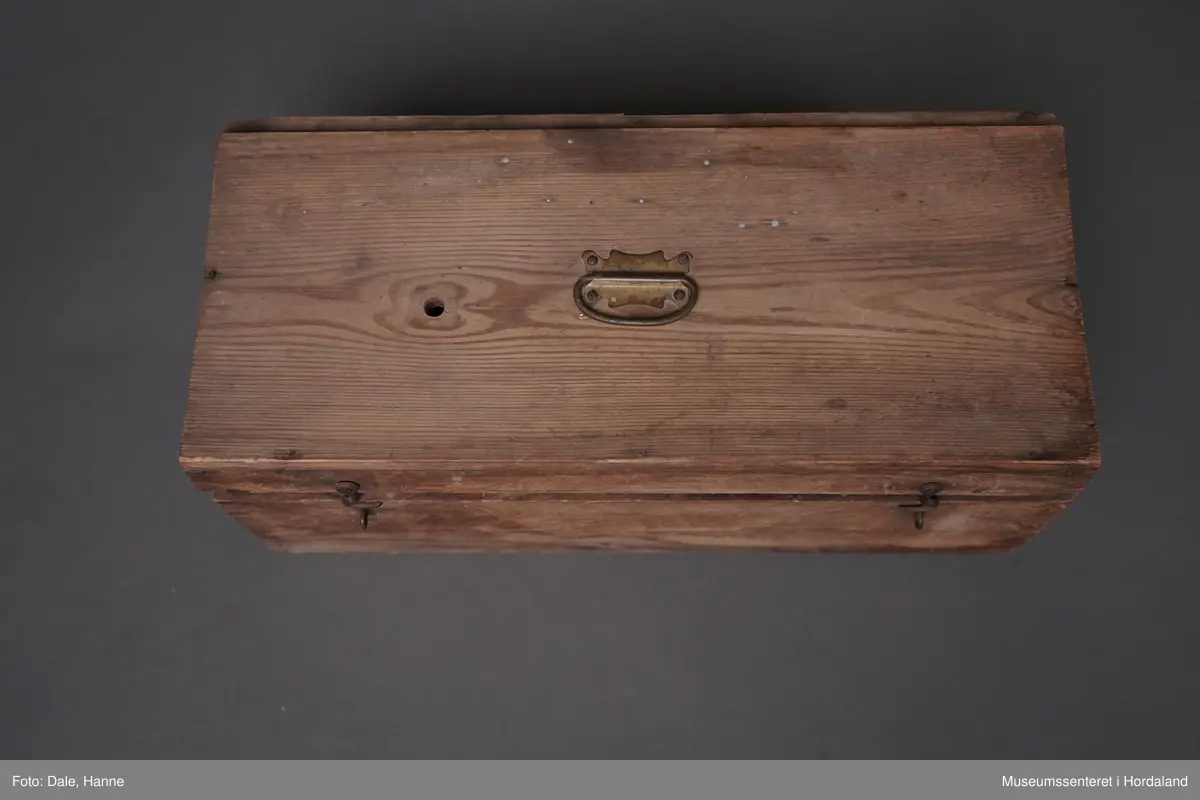 Svenskprodusert manuell rundstrikkemaskin med sveiv i oppbevaringskasse av tre. Det følgjer også med tre lodd og to trespolar.