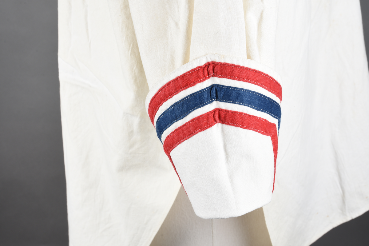 Matrosbluse av hvitt lerret. Matroskrave med to røde og en blå stripe. Lange ermer. Mansjetter som kraven. Rynket rygg fra bærestykke. knappes foran.
