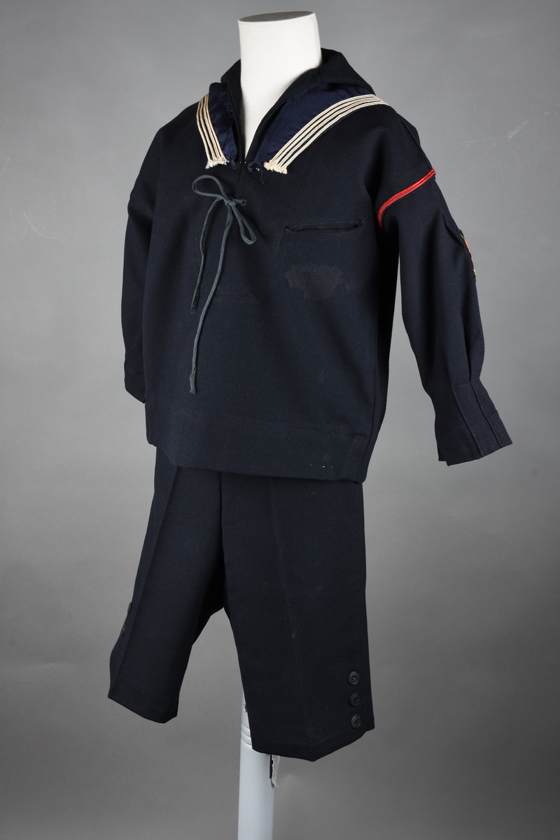 Matrosdress av ull og bomull. Bluse (A), benklær (B), livstykke (C) og matroskrave (D). Bluse med rett form og snøring i halsen. Legg på ermene. Lomme med rødt lommetørkle. Rød snor om skulder, Flagg- og våpenmerke. Benklær/bukse er åpen med knapp i begge sidene. Knelengde. Tre knappper på hvert bein, seks oventil. Grått livstykke til å feste buksen på. Knapphull og hemper. Matroskrave av mørkeblå silke med fire hvite striper langs den ytre kanten. To sorte bendel-knytebånd.