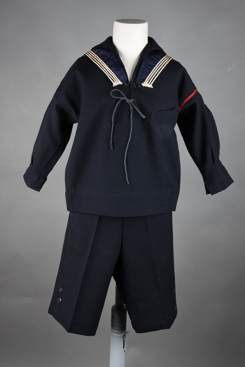 Matrosdress av ull og bomull. Bluse (A), benklær (B), livstykke (C) og matroskrave (D). Bluse med rett form og snøring i halsen. Legg på ermene. Lomme med rødt lommetørkle. Rød snor om skulder, Flagg- og våpenmerke. Benklær/bukse er åpen med knapp i begge sidene. Knelengde. Tre knappper på hvert bein, seks oventil. Grått livstykke til å feste buksen på. Knapphull og hemper. Matroskrave av mørkeblå silke med fire hvite striper langs den ytre kanten. To sorte bendel-knytebånd.