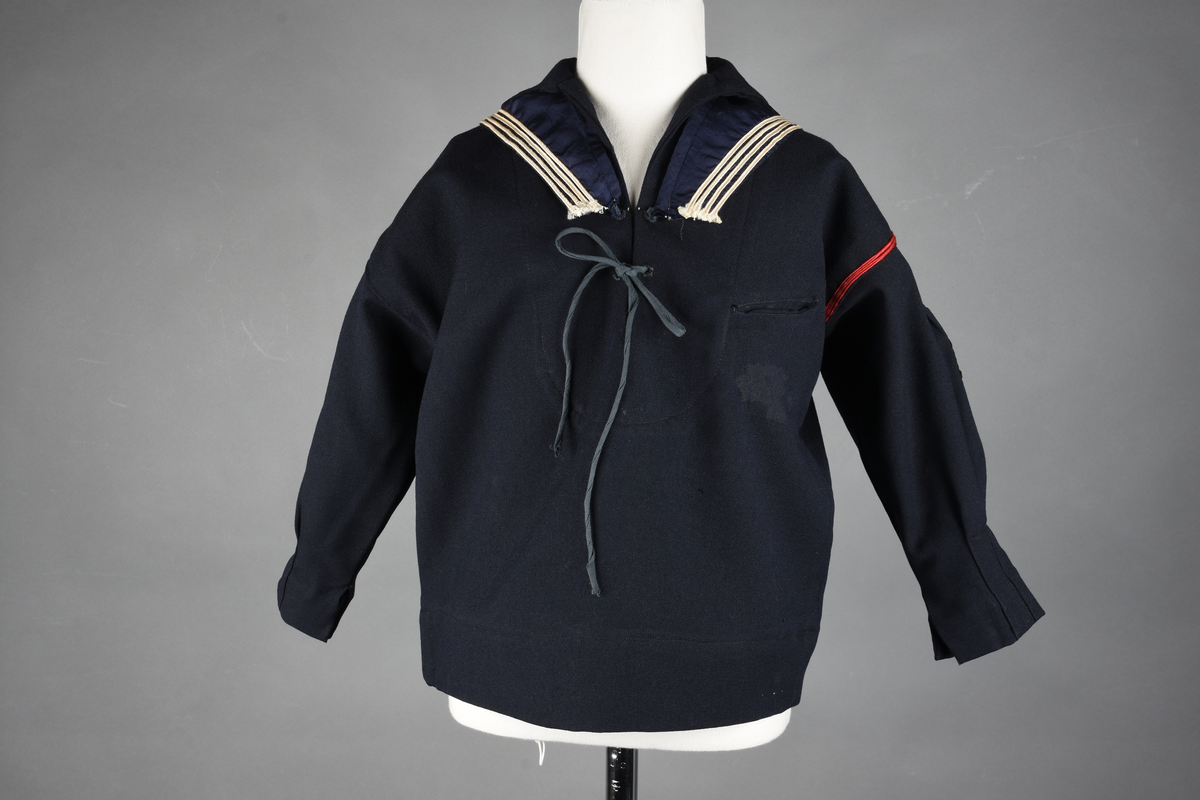 Matrosdress av ull og bomull. Bluse (A), benklær (B), livstykke (C) og matroskrave (D). Bluse med rett form og snøring i halsen. Legg på ermene. Lomme med rødt lommetørkle. Rød snor om skulder, Flagg- og våpenmerke. Benklær/bukse er åpen med knapp i begge sidene. Knelengde. Tre knappper på hvert bein, seks oventil. Grått livstykke til å feste buksen på. Knapphull og hemper. Matroskrave av mørkeblå silke med fire hvite striper langs den ytre kanten. To sorte bendel-knytebånd.