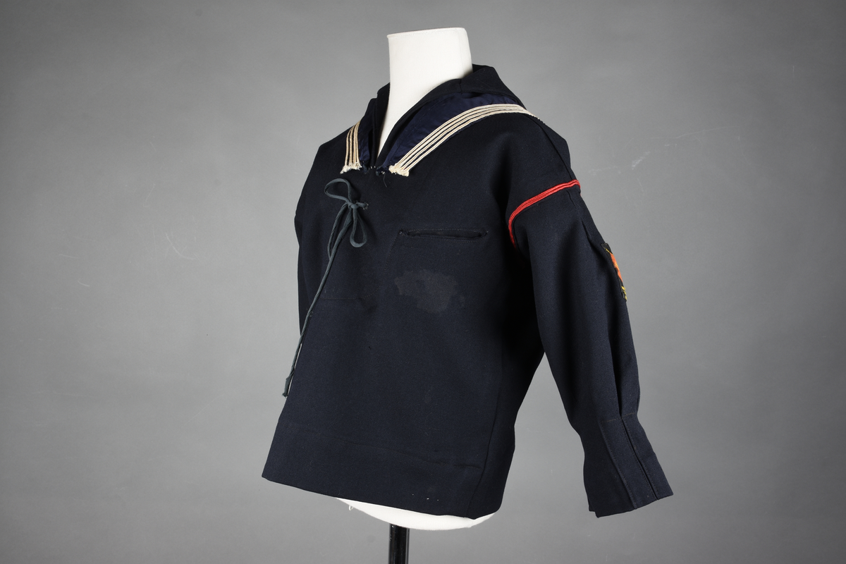 Matrosdress av ull og bomull. Bluse (A), benklær (B), livstykke (C) og matroskrave (D). Bluse med rett form og snøring i halsen. Legg på ermene. Lomme med rødt lommetørkle. Rød snor om skulder, Flagg- og våpenmerke. Benklær/bukse er åpen med knapp i begge sidene. Knelengde. Tre knappper på hvert bein, seks oventil. Grått livstykke til å feste buksen på. Knapphull og hemper. Matroskrave av mørkeblå silke med fire hvite striper langs den ytre kanten. To sorte bendel-knytebånd.