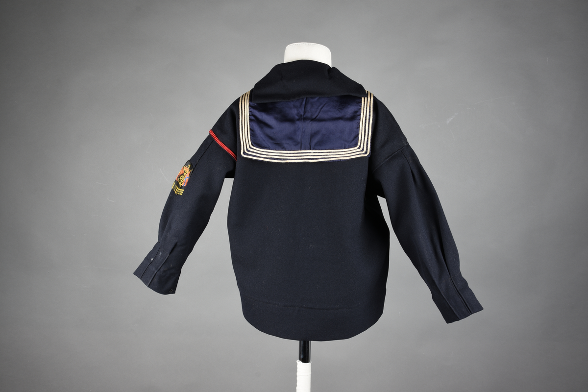 Matrosdress av ull og bomull. Bluse (A), benklær (B), livstykke (C) og matroskrave (D). Bluse med rett form og snøring i halsen. Legg på ermene. Lomme med rødt lommetørkle. Rød snor om skulder, Flagg- og våpenmerke. Benklær/bukse er åpen med knapp i begge sidene. Knelengde. Tre knappper på hvert bein, seks oventil. Grått livstykke til å feste buksen på. Knapphull og hemper. Matroskrave av mørkeblå silke med fire hvite striper langs den ytre kanten. To sorte bendel-knytebånd.