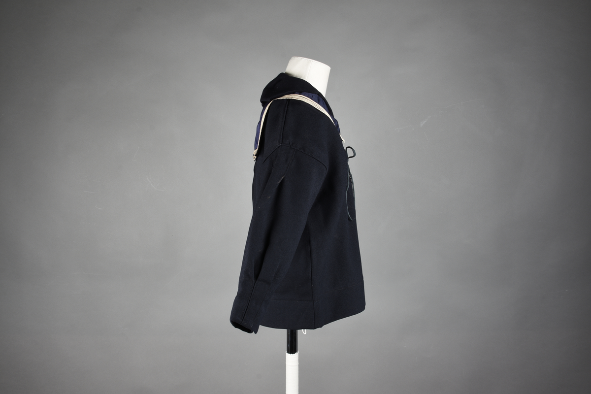 Matrosdress av ull og bomull. Bluse (A), benklær (B), livstykke (C) og matroskrave (D). Bluse med rett form og snøring i halsen. Legg på ermene. Lomme med rødt lommetørkle. Rød snor om skulder, Flagg- og våpenmerke. Benklær/bukse er åpen med knapp i begge sidene. Knelengde. Tre knappper på hvert bein, seks oventil. Grått livstykke til å feste buksen på. Knapphull og hemper. Matroskrave av mørkeblå silke med fire hvite striper langs den ytre kanten. To sorte bendel-knytebånd.