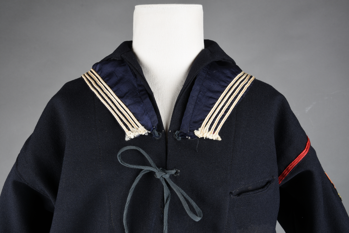 Matrosdress av ull og bomull. Bluse (A), benklær (B), livstykke (C) og matroskrave (D). Bluse med rett form og snøring i halsen. Legg på ermene. Lomme med rødt lommetørkle. Rød snor om skulder, Flagg- og våpenmerke. Benklær/bukse er åpen med knapp i begge sidene. Knelengde. Tre knappper på hvert bein, seks oventil. Grått livstykke til å feste buksen på. Knapphull og hemper. Matroskrave av mørkeblå silke med fire hvite striper langs den ytre kanten. To sorte bendel-knytebånd.