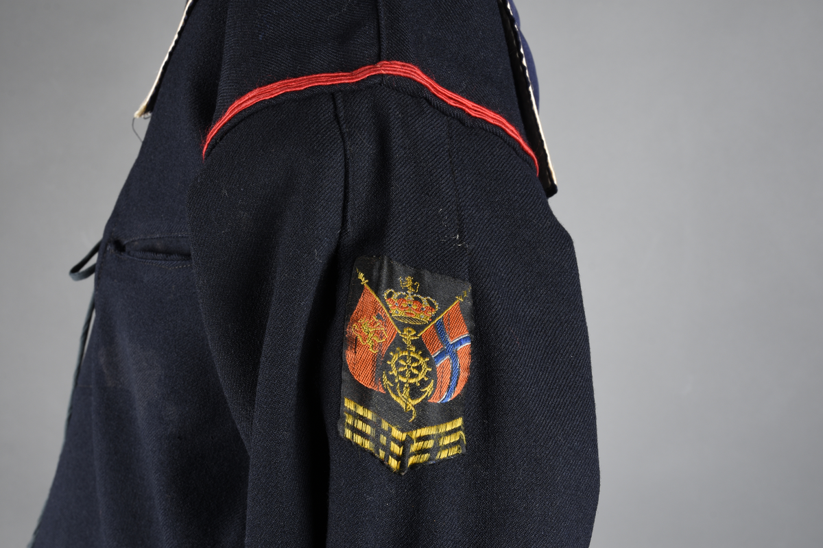 Matrosdress av ull og bomull. Bluse (A), benklær (B), livstykke (C) og matroskrave (D). Bluse med rett form og snøring i halsen. Legg på ermene. Lomme med rødt lommetørkle. Rød snor om skulder, Flagg- og våpenmerke. Benklær/bukse er åpen med knapp i begge sidene. Knelengde. Tre knappper på hvert bein, seks oventil. Grått livstykke til å feste buksen på. Knapphull og hemper. Matroskrave av mørkeblå silke med fire hvite striper langs den ytre kanten. To sorte bendel-knytebånd.