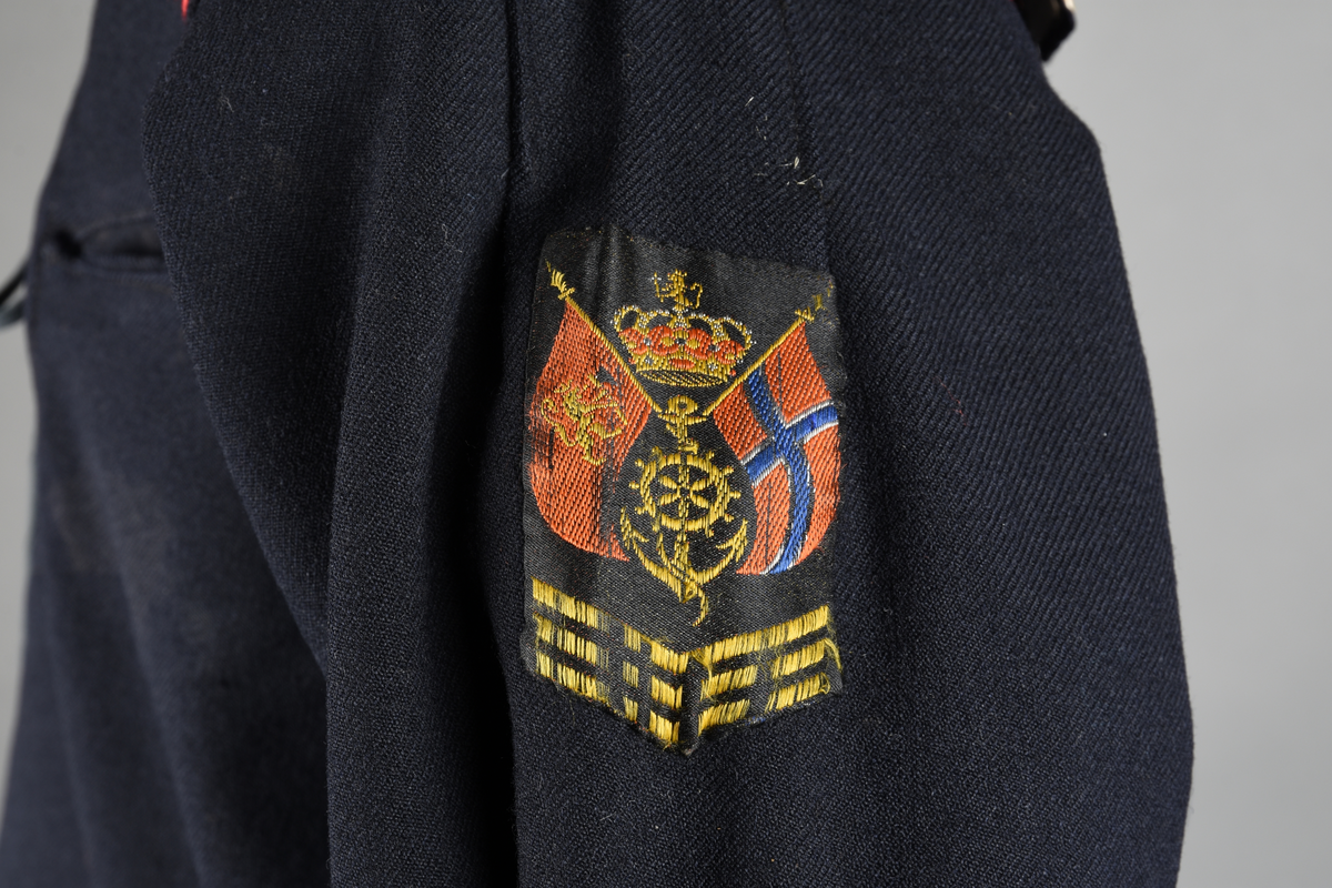 Matrosdress av ull og bomull. Bluse (A), benklær (B), livstykke (C) og matroskrave (D). Bluse med rett form og snøring i halsen. Legg på ermene. Lomme med rødt lommetørkle. Rød snor om skulder, Flagg- og våpenmerke. Benklær/bukse er åpen med knapp i begge sidene. Knelengde. Tre knappper på hvert bein, seks oventil. Grått livstykke til å feste buksen på. Knapphull og hemper. Matroskrave av mørkeblå silke med fire hvite striper langs den ytre kanten. To sorte bendel-knytebånd.