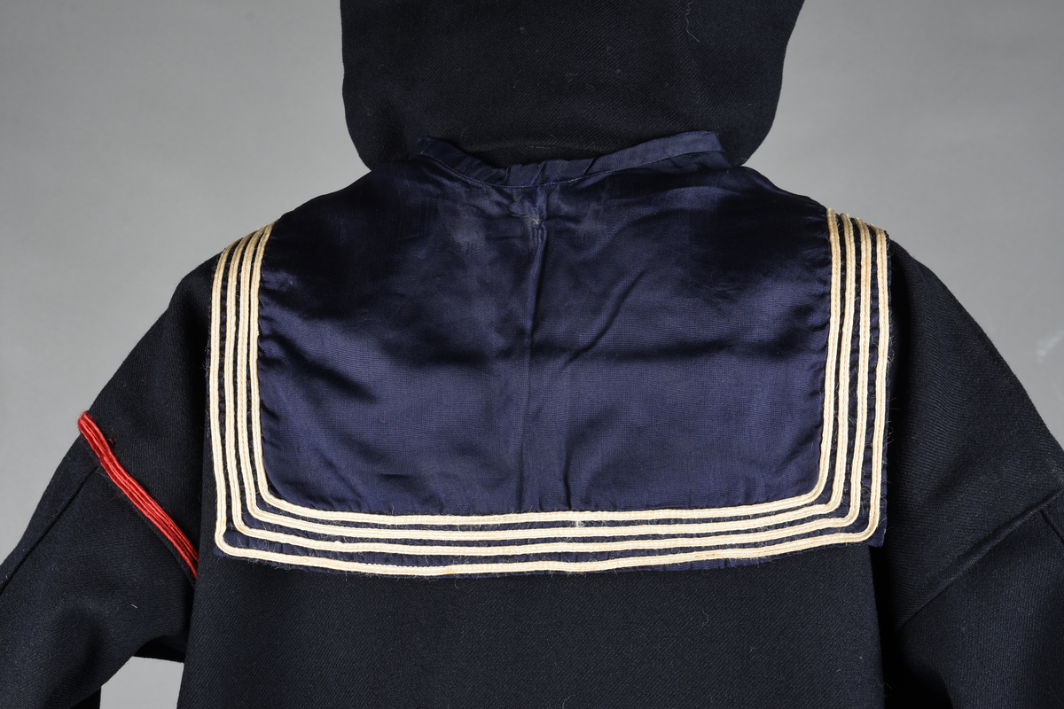 Matrosdress av ull og bomull. Bluse (A), benklær (B), livstykke (C) og matroskrave (D). Bluse med rett form og snøring i halsen. Legg på ermene. Lomme med rødt lommetørkle. Rød snor om skulder, Flagg- og våpenmerke. Benklær/bukse er åpen med knapp i begge sidene. Knelengde. Tre knappper på hvert bein, seks oventil. Grått livstykke til å feste buksen på. Knapphull og hemper. Matroskrave av mørkeblå silke med fire hvite striper langs den ytre kanten. To sorte bendel-knytebånd.