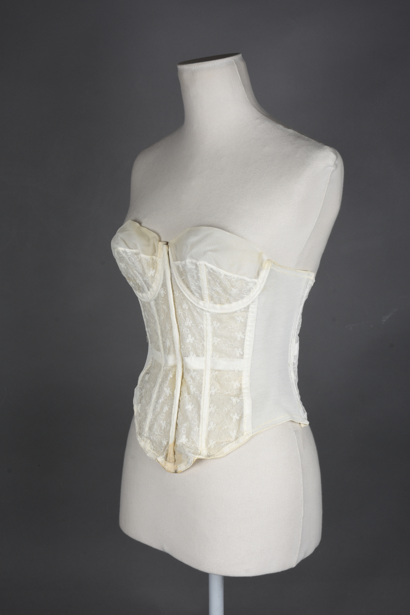 Brystholder av blondestrikk, med spiler. Både korsettspiler og cup-spiler. Lang form. Lukkes bak med hekter.