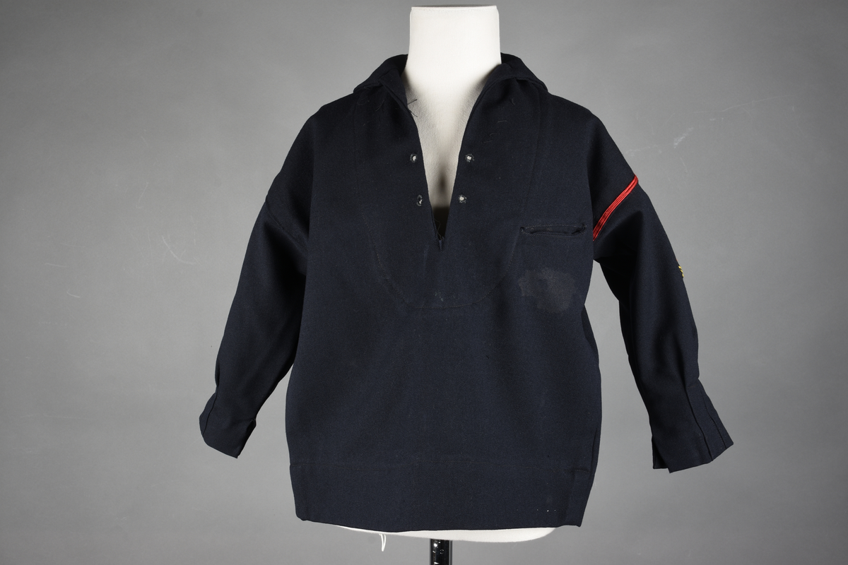 Matrosdress av ull og bomull. Bluse (A), benklær (B), livstykke (C) og matroskrave (D). Bluse med rett form og snøring i halsen. Legg på ermene. Lomme med rødt lommetørkle. Rød snor om skulder, Flagg- og våpenmerke. Benklær/bukse er åpen med knapp i begge sidene. Knelengde. Tre knappper på hvert bein, seks oventil. Grått livstykke til å feste buksen på. Knapphull og hemper. Matroskrave av mørkeblå silke med fire hvite striper langs den ytre kanten. To sorte bendel-knytebånd.