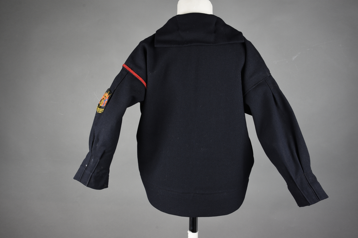 Matrosdress av ull og bomull. Bluse (A), benklær (B), livstykke (C) og matroskrave (D). Bluse med rett form og snøring i halsen. Legg på ermene. Lomme med rødt lommetørkle. Rød snor om skulder, Flagg- og våpenmerke. Benklær/bukse er åpen med knapp i begge sidene. Knelengde. Tre knappper på hvert bein, seks oventil. Grått livstykke til å feste buksen på. Knapphull og hemper. Matroskrave av mørkeblå silke med fire hvite striper langs den ytre kanten. To sorte bendel-knytebånd.