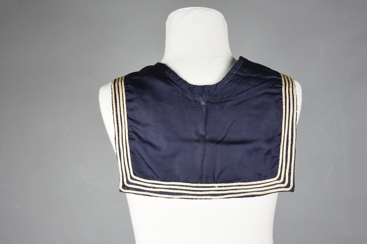 Matrosdress av ull og bomull. Bluse (A), benklær (B), livstykke (C) og matroskrave (D). Bluse med rett form og snøring i halsen. Legg på ermene. Lomme med rødt lommetørkle. Rød snor om skulder, Flagg- og våpenmerke. Benklær/bukse er åpen med knapp i begge sidene. Knelengde. Tre knappper på hvert bein, seks oventil. Grått livstykke til å feste buksen på. Knapphull og hemper. Matroskrave av mørkeblå silke med fire hvite striper langs den ytre kanten. To sorte bendel-knytebånd.