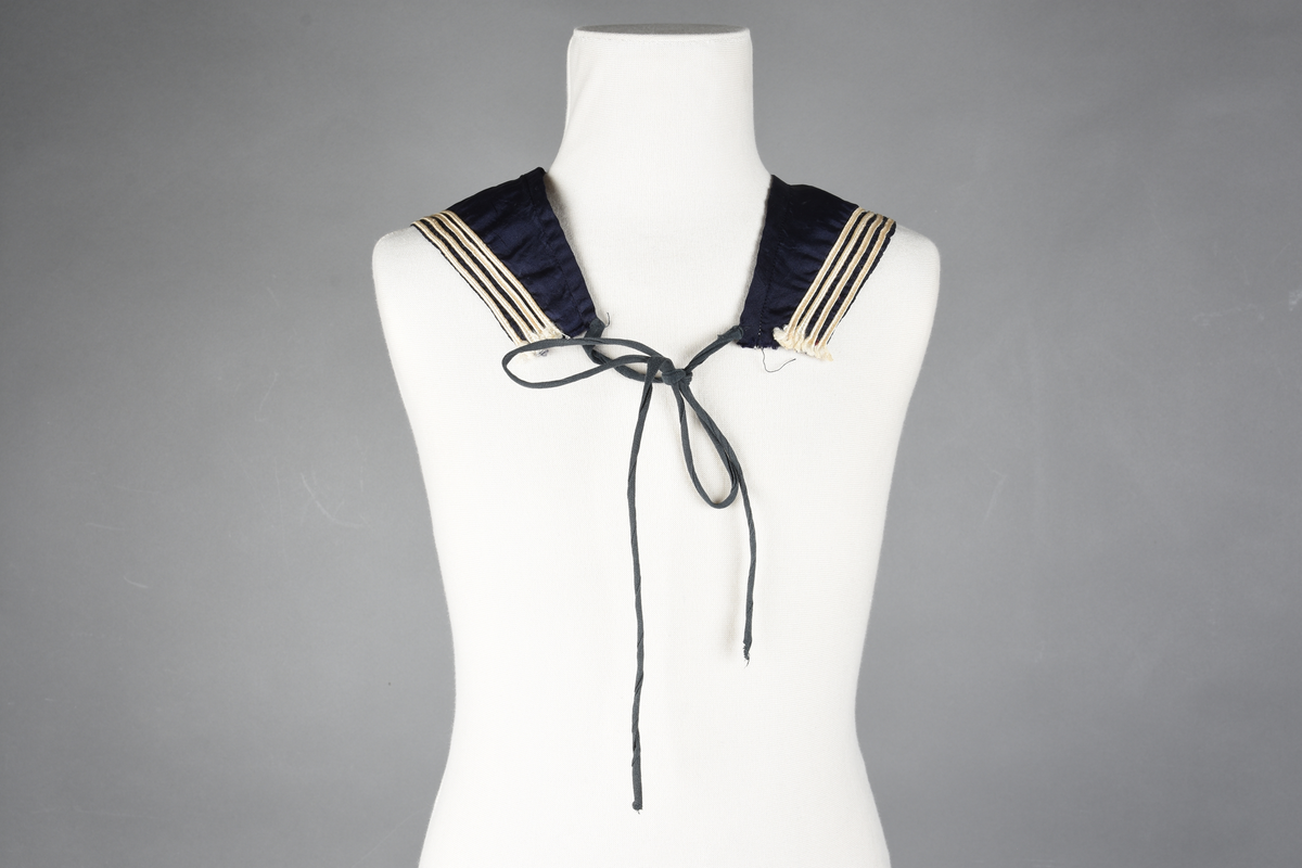 Matrosdress av ull og bomull. Bluse (A), benklær (B), livstykke (C) og matroskrave (D). Bluse med rett form og snøring i halsen. Legg på ermene. Lomme med rødt lommetørkle. Rød snor om skulder, Flagg- og våpenmerke. Benklær/bukse er åpen med knapp i begge sidene. Knelengde. Tre knappper på hvert bein, seks oventil. Grått livstykke til å feste buksen på. Knapphull og hemper. Matroskrave av mørkeblå silke med fire hvite striper langs den ytre kanten. To sorte bendel-knytebånd.