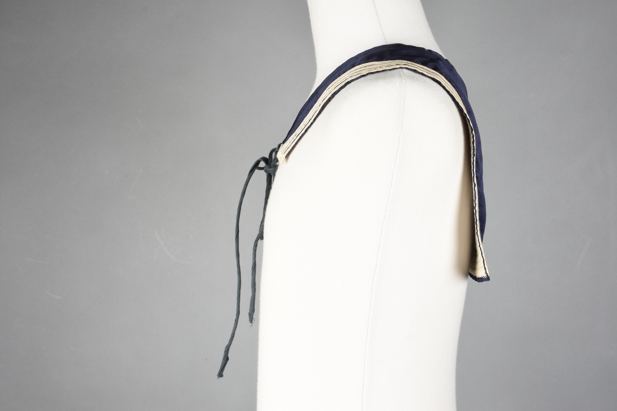 Matrosdress av ull og bomull. Bluse (A), benklær (B), livstykke (C) og matroskrave (D). Bluse med rett form og snøring i halsen. Legg på ermene. Lomme med rødt lommetørkle. Rød snor om skulder, Flagg- og våpenmerke. Benklær/bukse er åpen med knapp i begge sidene. Knelengde. Tre knappper på hvert bein, seks oventil. Grått livstykke til å feste buksen på. Knapphull og hemper. Matroskrave av mørkeblå silke med fire hvite striper langs den ytre kanten. To sorte bendel-knytebånd.