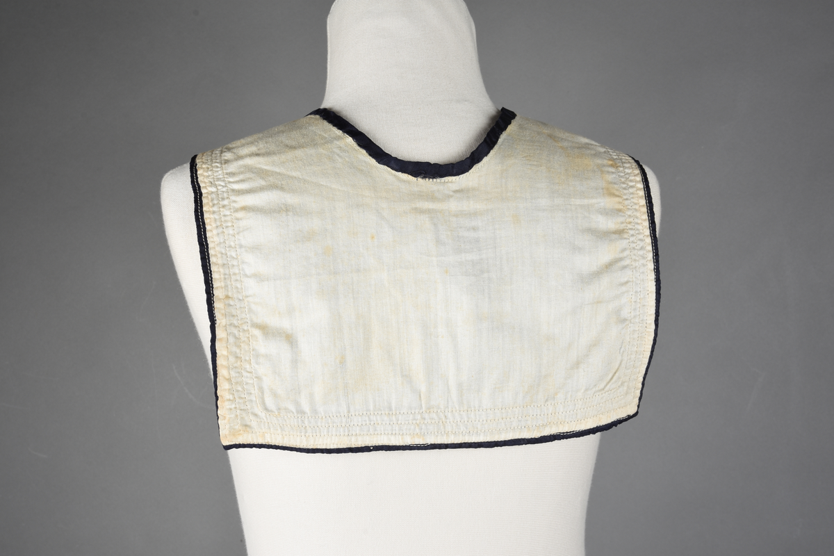 Matrosdress av ull og bomull. Bluse (A), benklær (B), livstykke (C) og matroskrave (D). Bluse med rett form og snøring i halsen. Legg på ermene. Lomme med rødt lommetørkle. Rød snor om skulder, Flagg- og våpenmerke. Benklær/bukse er åpen med knapp i begge sidene. Knelengde. Tre knappper på hvert bein, seks oventil. Grått livstykke til å feste buksen på. Knapphull og hemper. Matroskrave av mørkeblå silke med fire hvite striper langs den ytre kanten. To sorte bendel-knytebånd.