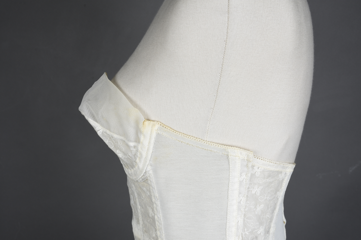 Brystholder av blondestrikk, med spiler. Både korsettspiler og cup-spiler. Lang form. Lukkes bak med hekter.