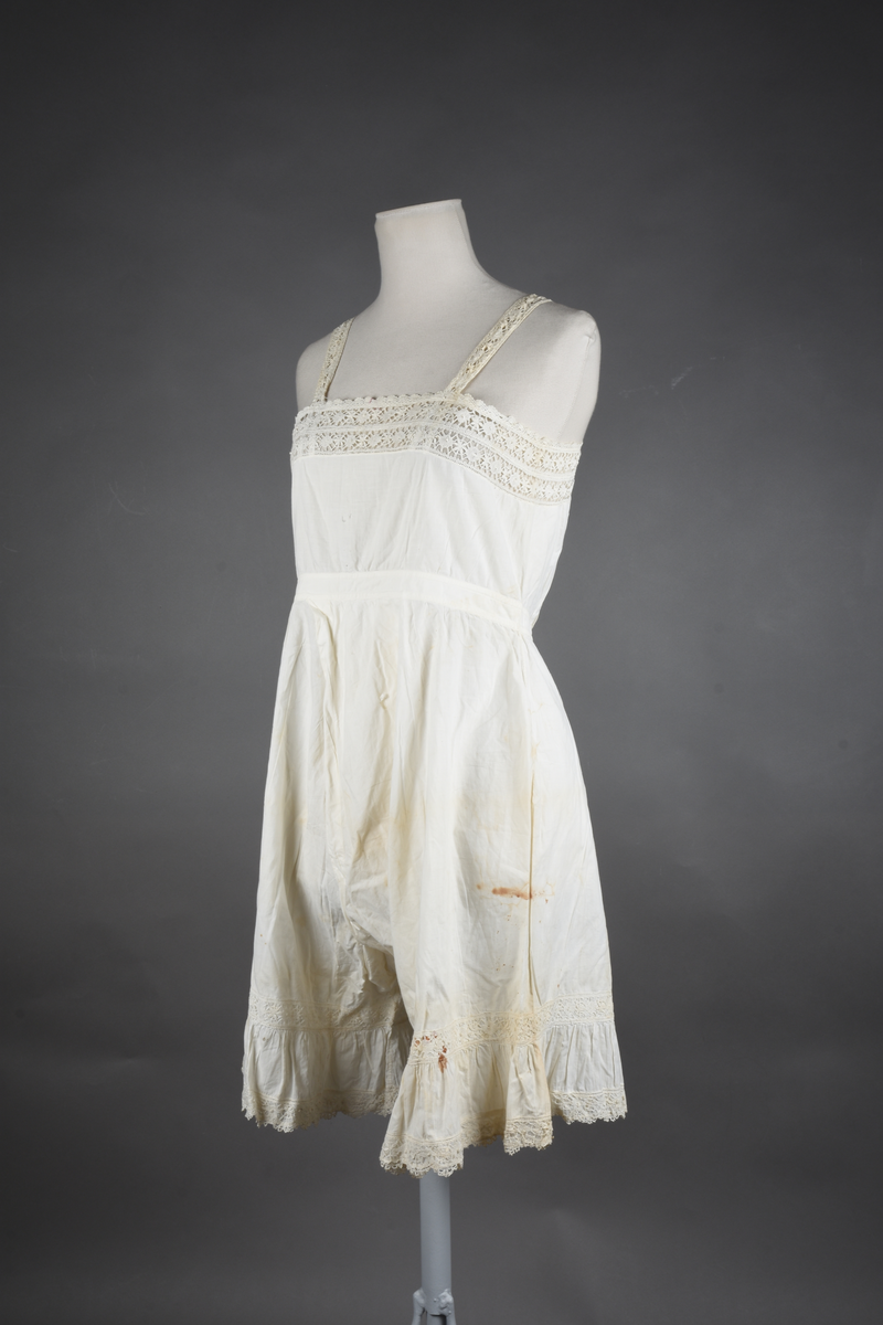 Kombinasjon av fint, hvitt bomullslerret. Livet har smale blonde-skulderstropper festet mot en bred blondekant. Foran er kombinasjonen rynket til en linning i taillen. Benklærne er sydd til samme linning og de har lavt skritt og er helt åpne nedentil. Nederst er de kantet med rynkekappe og blonder. Knappet foran med ti små knapper. Noen rustflekker. Lite hull revet bak.