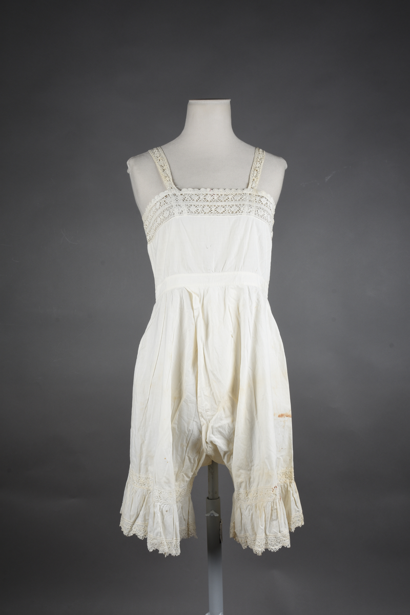 Kombinasjon av fint, hvitt bomullslerret. Livet har smale blonde-skulderstropper festet mot en bred blondekant. Foran er kombinasjonen rynket til en linning i taillen. Benklærne er sydd til samme linning og de har lavt skritt og er helt åpne nedentil. Nederst er de kantet med rynkekappe og blonder. Knappet foran med ti små knapper. Noen rustflekker. Lite hull revet bak.