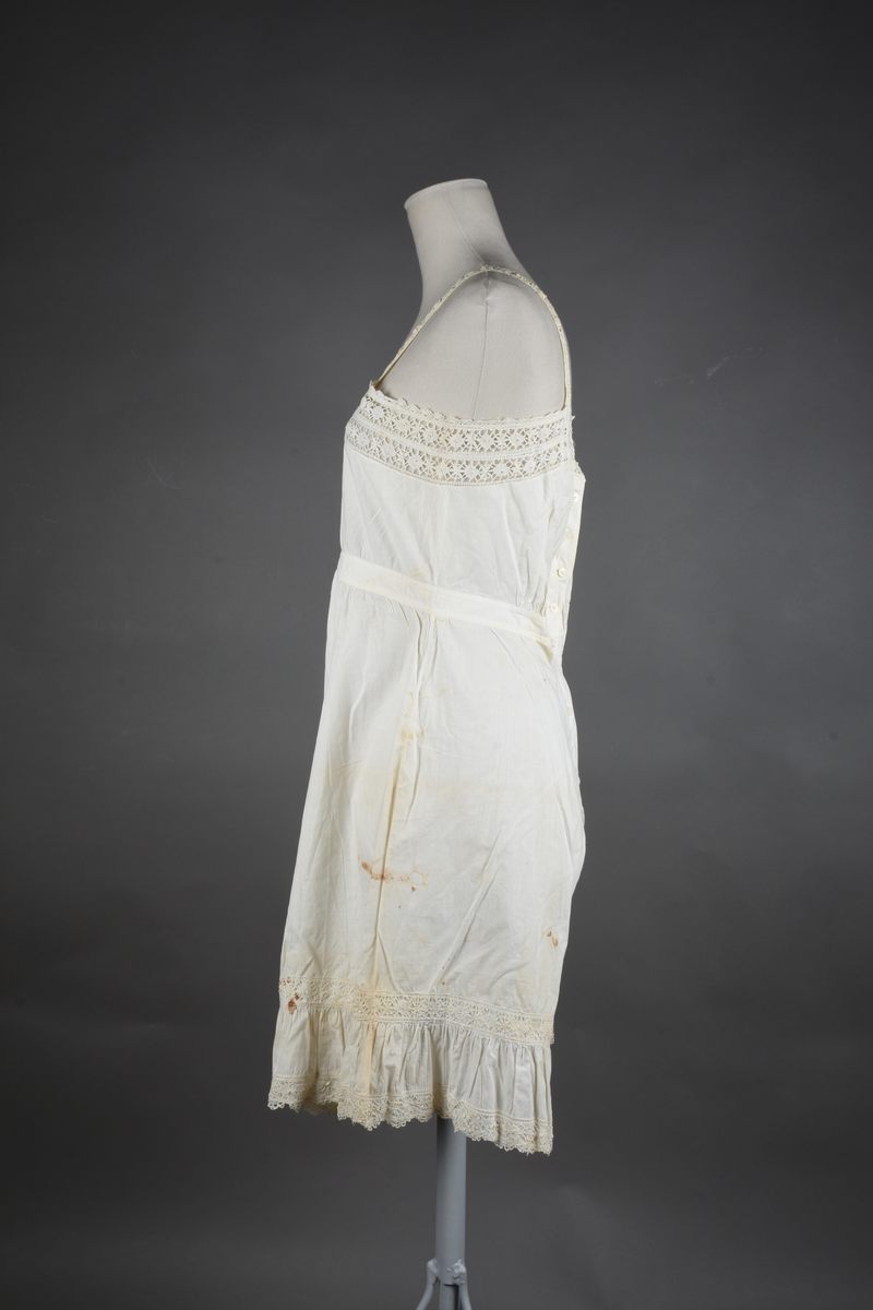 Kombinasjon av fint, hvitt bomullslerret. Livet har smale blonde-skulderstropper festet mot en bred blondekant. Foran er kombinasjonen rynket til en linning i taillen. Benklærne er sydd til samme linning og de har lavt skritt og er helt åpne nedentil. Nederst er de kantet med rynkekappe og blonder. Knappet foran med ti små knapper. Noen rustflekker. Lite hull revet bak.