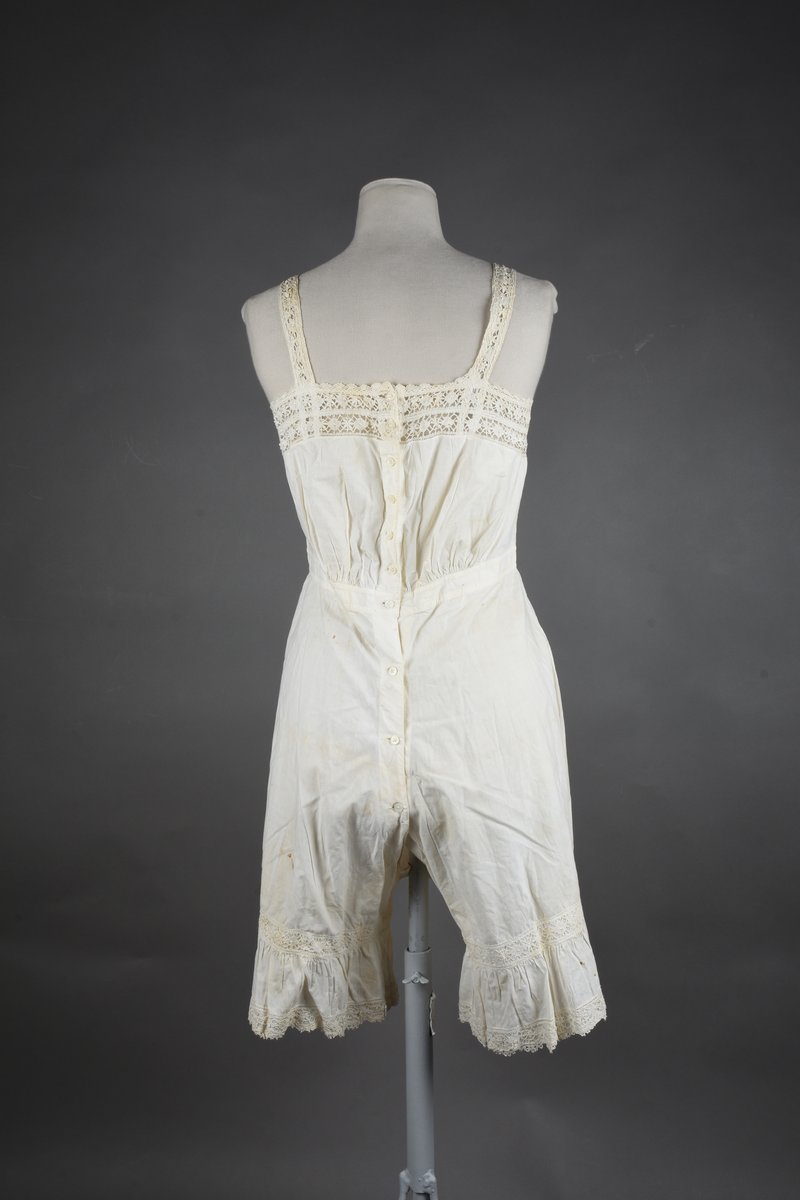 Kombinasjon av fint, hvitt bomullslerret. Livet har smale blonde-skulderstropper festet mot en bred blondekant. Foran er kombinasjonen rynket til en linning i taillen. Benklærne er sydd til samme linning og de har lavt skritt og er helt åpne nedentil. Nederst er de kantet med rynkekappe og blonder. Knappet foran med ti små knapper. Noen rustflekker. Lite hull revet bak.