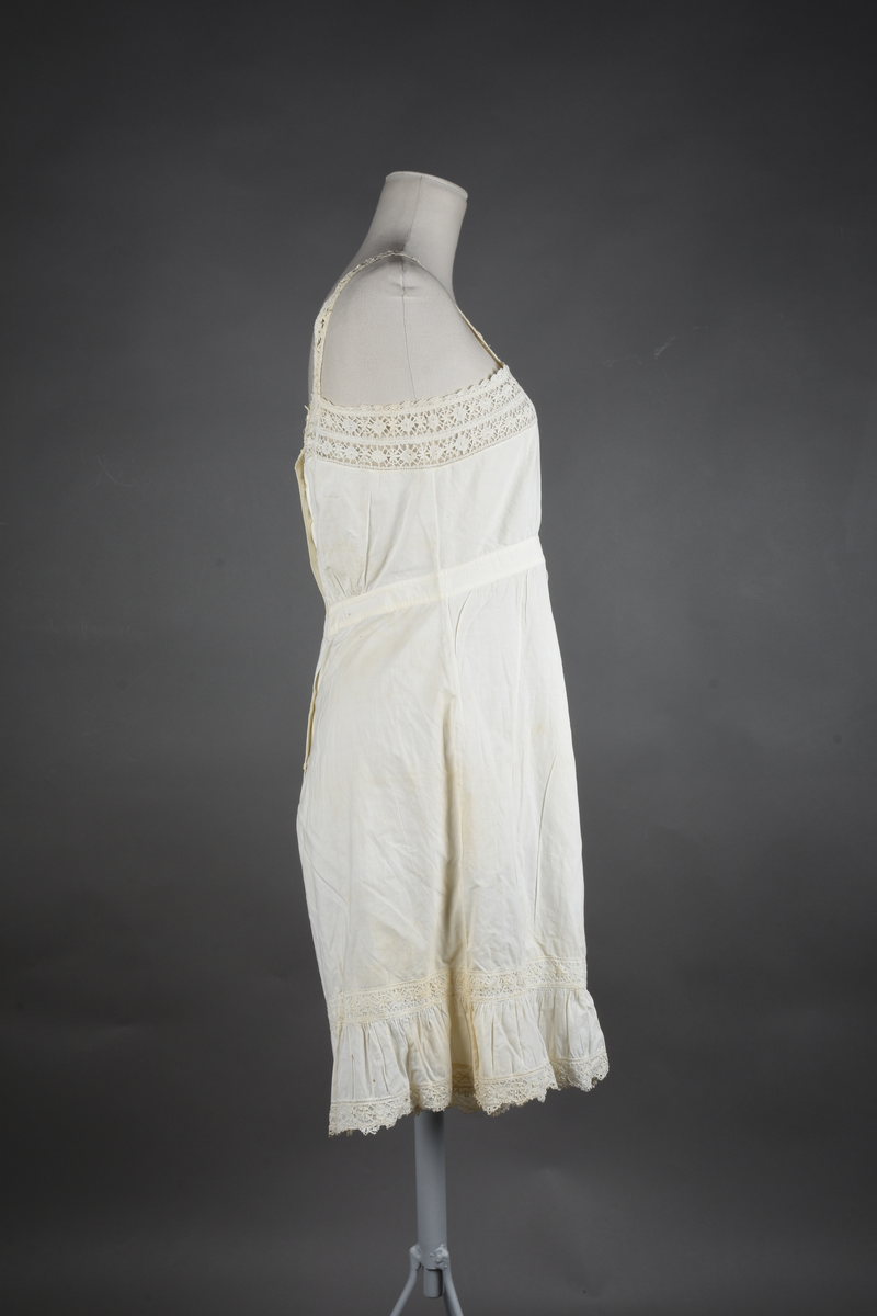 Kombinasjon av fint, hvitt bomullslerret. Livet har smale blonde-skulderstropper festet mot en bred blondekant. Foran er kombinasjonen rynket til en linning i taillen. Benklærne er sydd til samme linning og de har lavt skritt og er helt åpne nedentil. Nederst er de kantet med rynkekappe og blonder. Knappet foran med ti små knapper. Noen rustflekker. Lite hull revet bak.