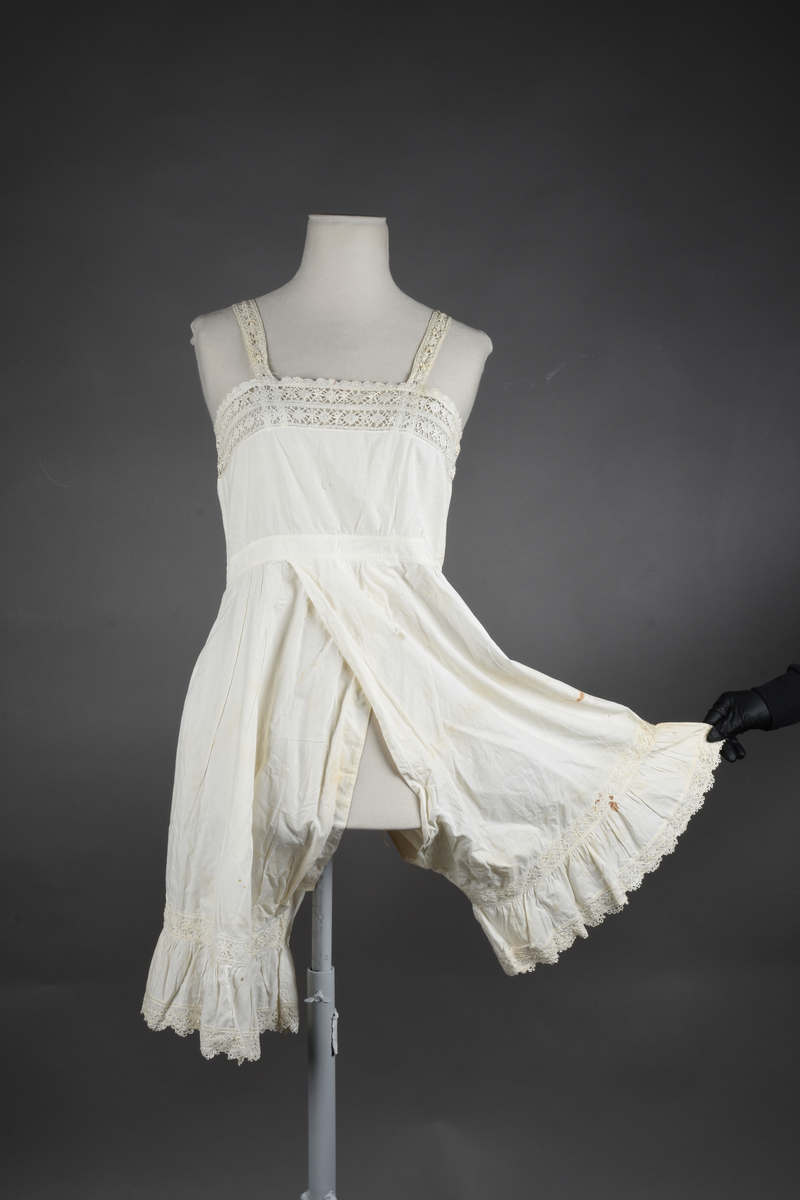 Kombinasjon av fint, hvitt bomullslerret. Livet har smale blonde-skulderstropper festet mot en bred blondekant. Foran er kombinasjonen rynket til en linning i taillen. Benklærne er sydd til samme linning og de har lavt skritt og er helt åpne nedentil. Nederst er de kantet med rynkekappe og blonder. Knappet foran med ti små knapper. Noen rustflekker. Lite hull revet bak.