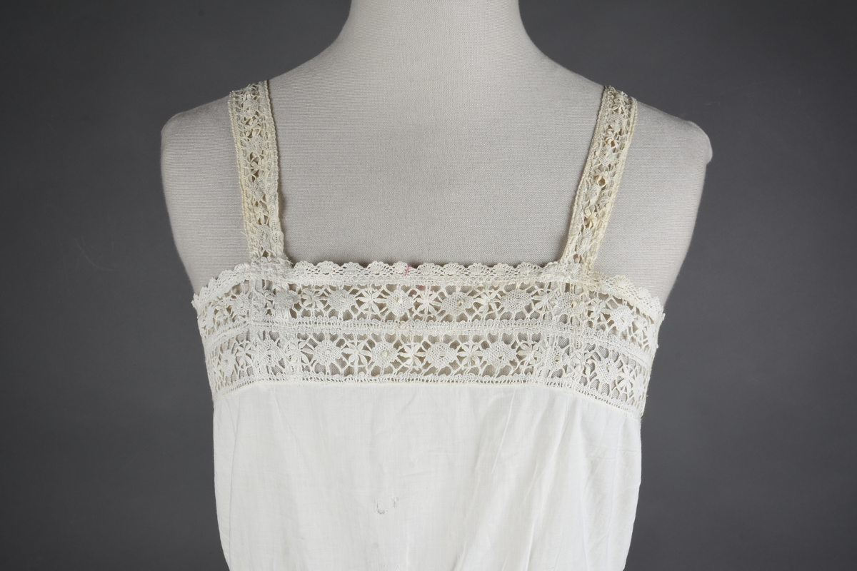 Kombinasjon av fint, hvitt bomullslerret. Livet har smale blonde-skulderstropper festet mot en bred blondekant. Foran er kombinasjonen rynket til en linning i taillen. Benklærne er sydd til samme linning og de har lavt skritt og er helt åpne nedentil. Nederst er de kantet med rynkekappe og blonder. Knappet foran med ti små knapper. Noen rustflekker. Lite hull revet bak.