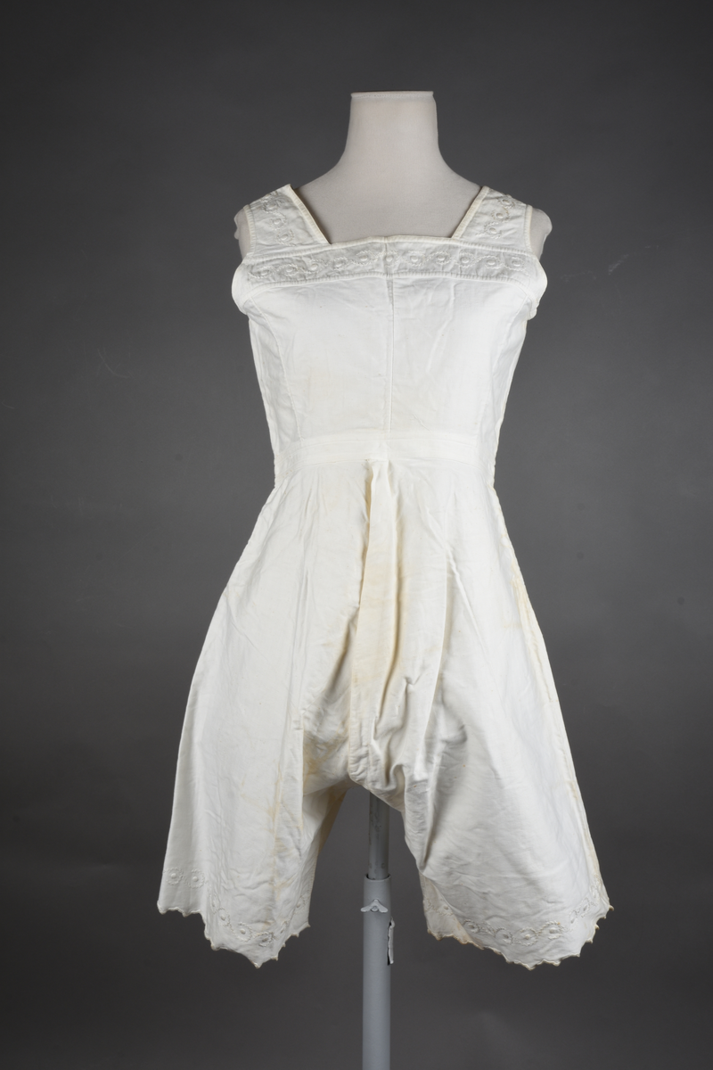 Kombinasjon av bomullslerret. Kraftig kypervevd lerret (twill). Overdelen har brede skulderstropper og er figursydd til en linning i taillen. Benklærne er sydd til samme linning og de har lavt skritt og er helt åpne nedentil. Knappet foran med elleve små knapper. Broderi og tungekant nederst - “løpende hund”-ranke rundt store prikker, tungekant på beina. Noen rustflekker. Lite hull revet bak.