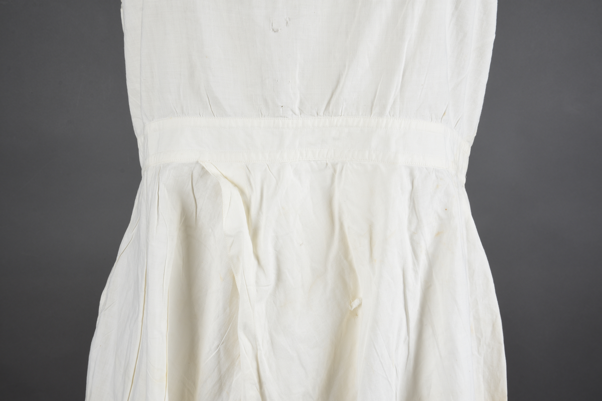 Kombinasjon av fint, hvitt bomullslerret. Livet har smale blonde-skulderstropper festet mot en bred blondekant. Foran er kombinasjonen rynket til en linning i taillen. Benklærne er sydd til samme linning og de har lavt skritt og er helt åpne nedentil. Nederst er de kantet med rynkekappe og blonder. Knappet foran med ti små knapper. Noen rustflekker. Lite hull revet bak.