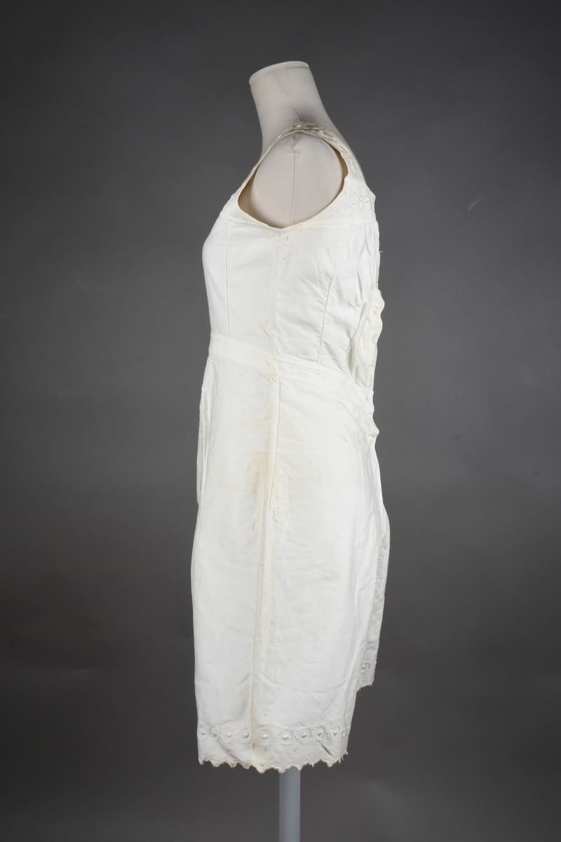 Kombinasjon av bomullslerret. Kraftig kypervevd lerret (twill). Overdelen har brede skulderstropper og er figursydd til en linning i taillen. Benklærne er sydd til samme linning og de har lavt skritt og er helt åpne nedentil. Knappet foran med elleve små knapper. Broderi og tungekant nederst - “løpende hund”-ranke rundt store prikker, tungekant på beina. Noen rustflekker. Lite hull revet bak.