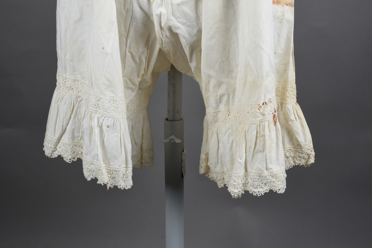 Kombinasjon av fint, hvitt bomullslerret. Livet har smale blonde-skulderstropper festet mot en bred blondekant. Foran er kombinasjonen rynket til en linning i taillen. Benklærne er sydd til samme linning og de har lavt skritt og er helt åpne nedentil. Nederst er de kantet med rynkekappe og blonder. Knappet foran med ti små knapper. Noen rustflekker. Lite hull revet bak.