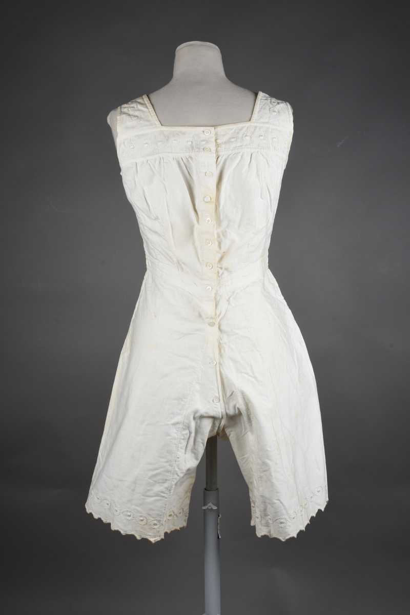 Kombinasjon av bomullslerret. Kraftig kypervevd lerret (twill). Overdelen har brede skulderstropper og er figursydd til en linning i taillen. Benklærne er sydd til samme linning og de har lavt skritt og er helt åpne nedentil. Knappet foran med elleve små knapper. Broderi og tungekant nederst - “løpende hund”-ranke rundt store prikker, tungekant på beina. Noen rustflekker. Lite hull revet bak.